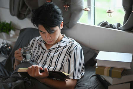 Peaceful young man drinking coffee and reading book on bed.