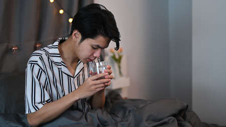 Asian man drinking water from a glass while sitting on his bed at night.の写真素材