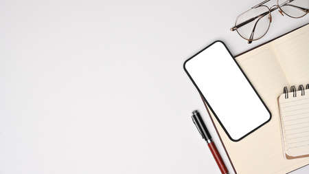 Mock up smart phone with blank screen, notebook and glasses on white office desk.の写真素材