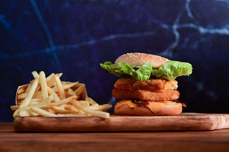 Homemade crispy fish burger with cheese, lettuce, and fried potatoes on wooden table.