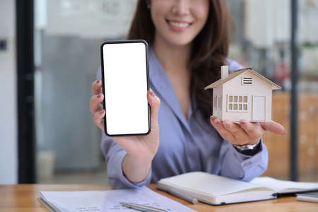Real estate agent woman showing smart phone with empty screen and holding house model.の写真素材