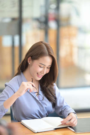 Portrait of confident businesswoman holding glasses and reading a book at office desk.の写真素材