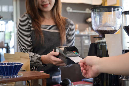 Female waiter holding credit card swipe machine while customer making payment in coffee shop with credit card.の写真素材