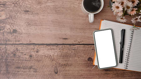 Top view mock up smart phone with white screen, coffee cup and notebook on wooden table,