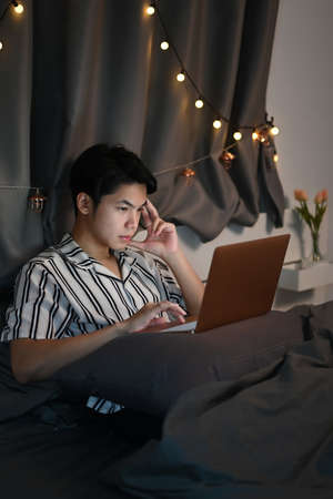 Young Asian man working with computer laptop on his bed at late night.
