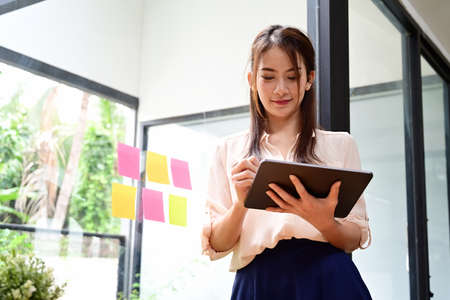 Confident businesswoman standing near glass window in office and using digital tablet.