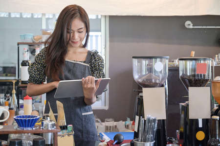 Smiling young Asian female entrepreneur in apron and using digital tablet while standing in her cafe.の写真素材