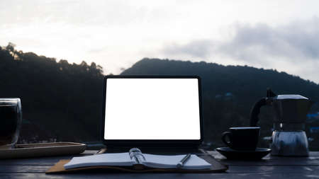 Computer tablet, notebook, moka pot and coffee cup on wooden table with mountain view in background.の写真素材