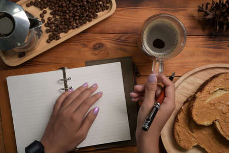 Woman holding glass of coffee and making notes on notebook.