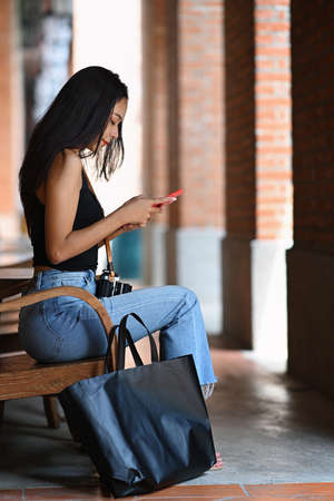 Smiling young woman using smart phone and sitting on stairs with shopping bags.の写真素材