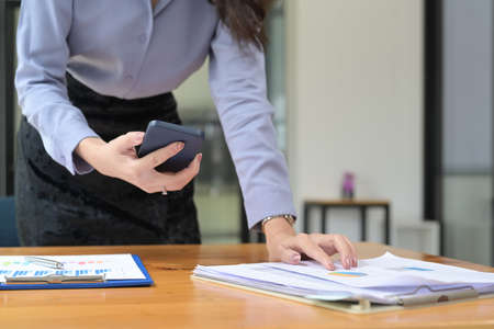 Cropped shot businesswoman holding smart phone and checking financial reports at office desk.