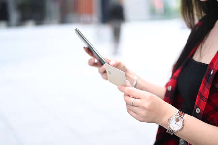 Young woman holding credit card and using smart phone for banking online while sitting at outdoor.の写真素材
