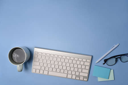 Top view wireless keyboard, coffee cup and glasses on blue background.