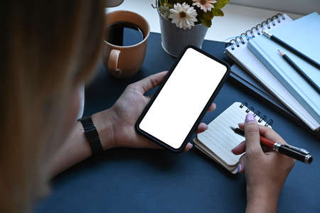 Young woman holding smart phone and making notes on notebook. Blank screen for advertise text.の写真素材