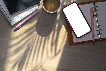 Mock up smart phone, coffee cup and notebook on wooden table near window.