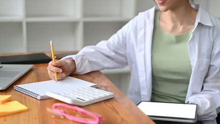 Cropped shot young woman holding digital tablet and writing information on notepad.