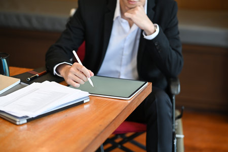 Disabled businessman sitting in wheelchair working with digital tablet at workplace.の写真素材