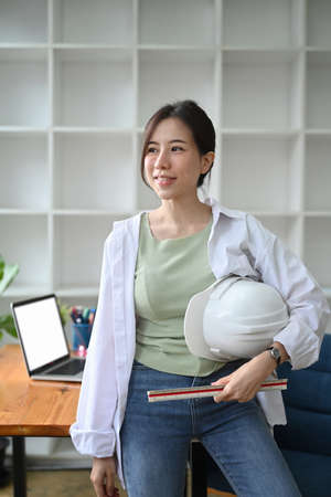Confident Architect woman holding with hard hat safety helmet and standing in office.の写真素材