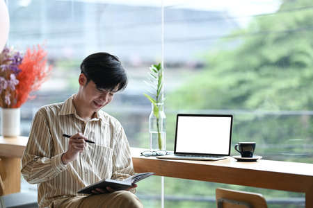 Smiling young man entrepreneur sitting in bright office and making notes on notebook.の写真素材