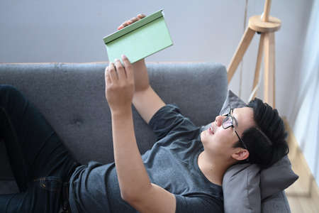 Happy Asian man laying on comfortable sofa and reading book.