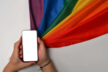 Male holding smart phone with blank screen near rainbow flag on white background.