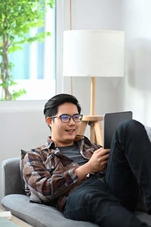 Happy man laying on sofa and checking social media with digital tablet.の写真素材