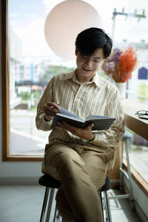 Smiling young Asian man sitting in bright office and reading book.
