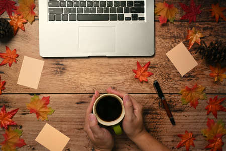 Overhead view man holding cup of hot beverage on wooden table with autumn maple leaves.の写真素材