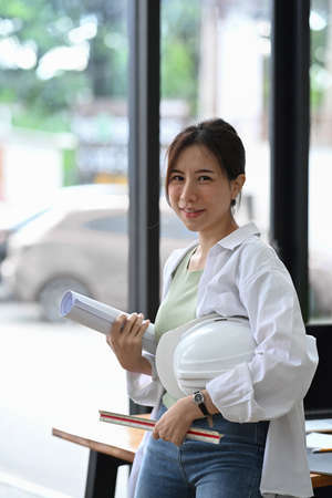 Beautiful engineer woman holding helmet and looking at camera while standing in office.の写真素材