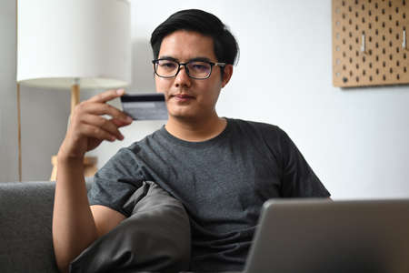 Handsome man holding credit card and using laptop computer in living room.の写真素材
