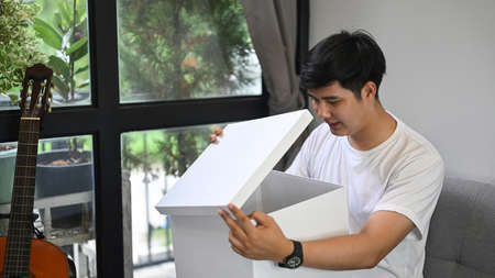 Happy man opening gift box while siting on sofa at home.の写真素材