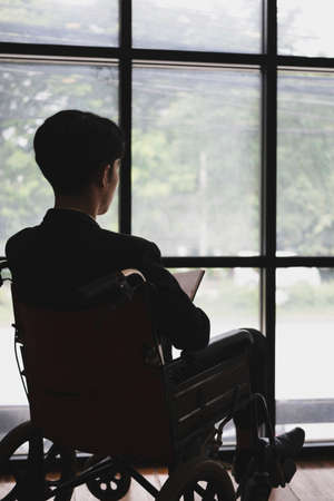 Rear view of thoughtful businessman in wheelchair sitting in office room and looking out of window.の写真素材