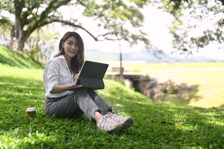 Happy woman sitting on grass with computer laptop in public park.の写真素材