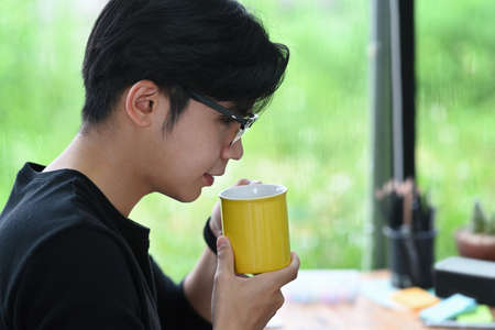 Peaceful young asian man sitting in office and drinking hot coffee.の写真素材