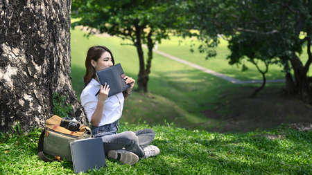 Peaceful young woman sitting under tree in the park and reading interesting book.の写真素材