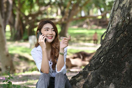 Peaceful young woman sitting under tree in the park and reading interesting book.の写真素材