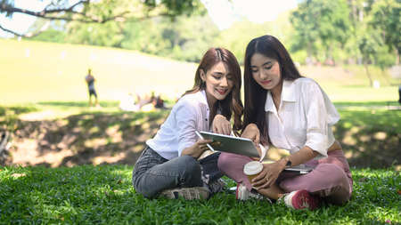 Two young asian women sitting on grass in the city park and working together with digital tablet.の写真素材