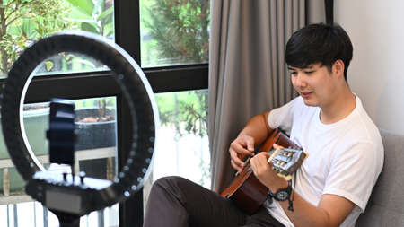 Young asian man video blogger playing guitar and recording video content for his blog.