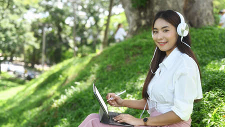 Asian woman wearing headphone and working with computer tablet in the park.