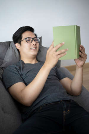 Smiling young asian man reading book on sofa.の写真素材
