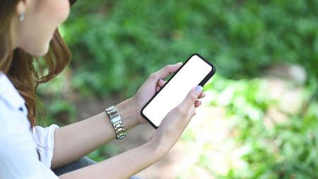 Cropped shot young woman holding mock up smart phone with blank screen while sitting in the park.の写真素材