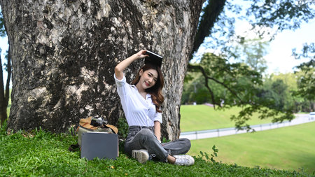 Beautiful woman relaxing and breathing fresh air while sitting on green grass under tree .
