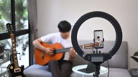 Young asian man playing guitar and streaming online vlog with smart phone at home.
