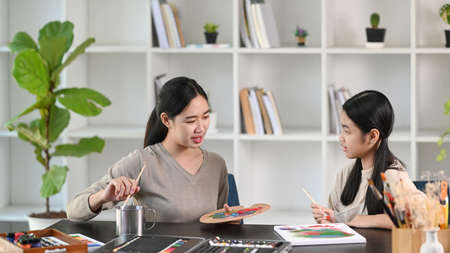 Smiling female teacher helping little girl painting picture in art class.