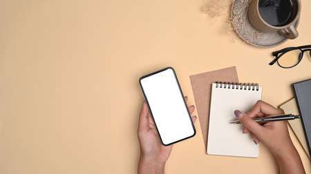 Overhead view female holding smart phone ad making notes on notebook.の写真素材