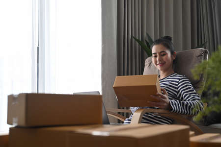 Asian woman small business entrepreneur holding cardboard box and checking orders on computer...