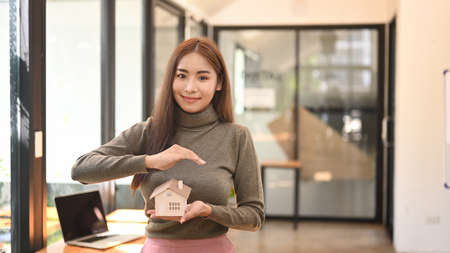 Young asian woman holding small home model  and smiling to camera. Real Estate Concept.