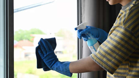 Cropped shot young man wearing rubber gloves cleaning window with rag and spray cleaner at home
