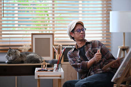 Asian man artist sitting in front of the canvas in living room and painting with water color.の写真素材