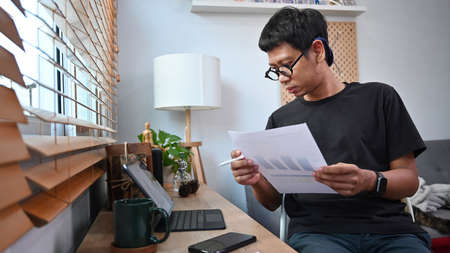 Young asian man holding financial graphs and statistics documents and working with computer tablet...